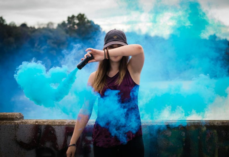 A stylish woman surrounded by bright blue smoke outdoors, creating a dynamic fashion statement.